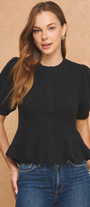 A woman modeling a black short-sleeve puff-sleeve top with a peplum hem, paired with blue jeans, against a textured tan background.