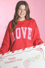 Woman wearing a red sweatshirt with 'LOVE' holding a Valentine's Day card against a pink background
