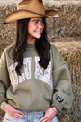 Woman wearing a green sweatshirt with Lulu Mac and a cowboy hat, standing in front of hay bales.