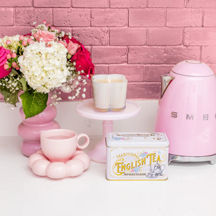 Pink tea set with a teapot, cup, and English tea box against a pink brick wall.