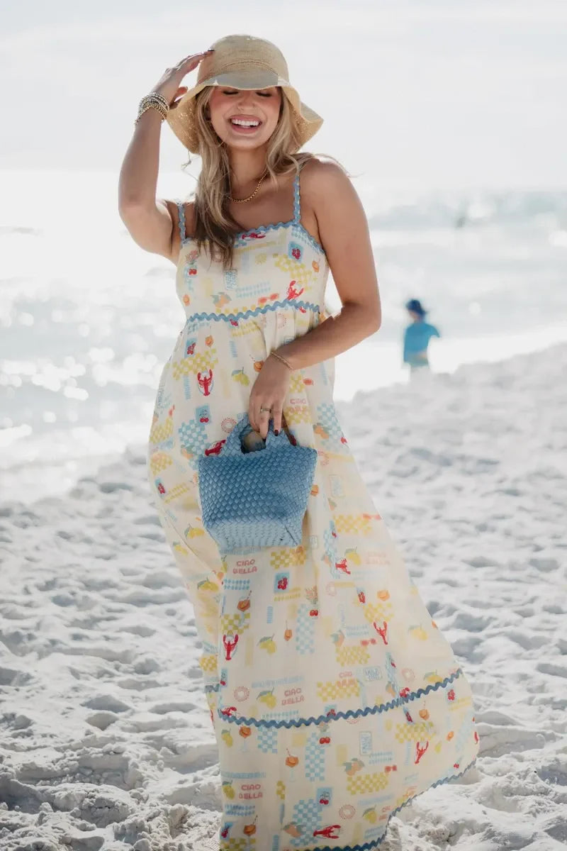 Woman in a long, patterned dress on a beach holding a blue bag.