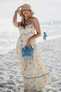 Woman in a long, patterned dress on a beach holding a blue bag.
