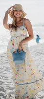 Woman in a long, patterned dress on a beach holding a blue bag.