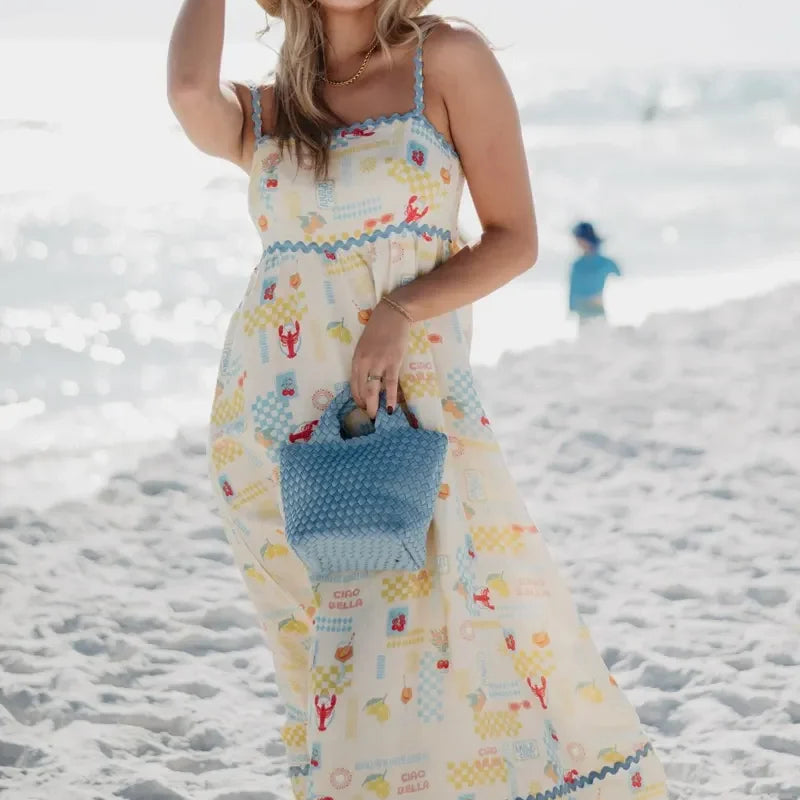 Woman in a long, patterned dress on a beach holding a blue bag.