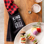 Black towel with 'King of the Grill' text on a wooden surface with skewered vegetables and drinks.