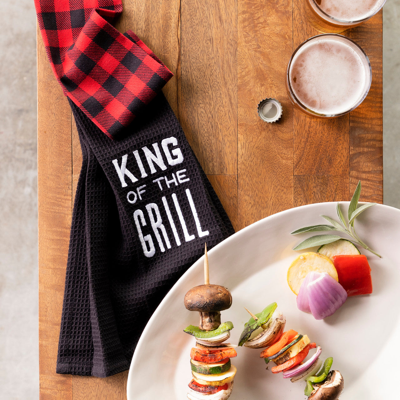 Black towel with 'King of the Grill' text on a wooden surface with skewered vegetables and drinks.