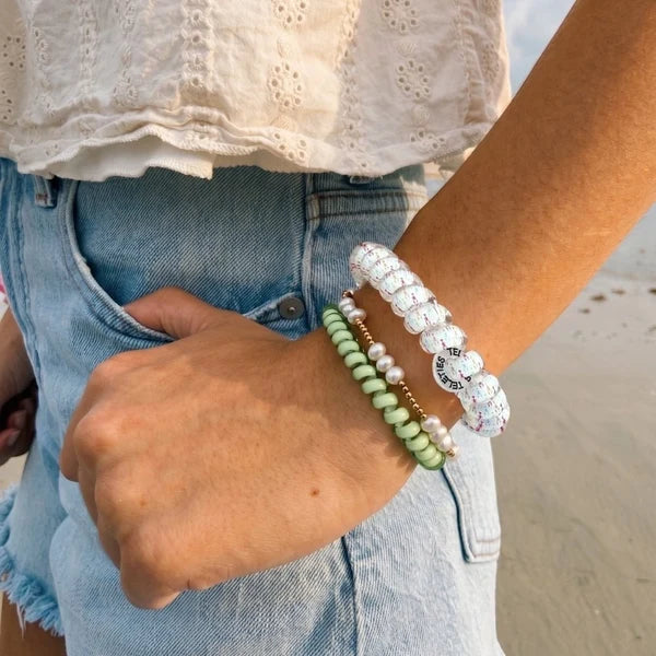 A close-up of a person's wrist wearing multiple beaded bracelets, including a white beaded bracelet with a silver moon charm, a mint green beaded bracelet, and a white pearl bracelet, paired with denim shorts.