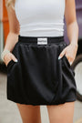 Person wearing a black skirt with a visible brand label, standing against a blurred background.
