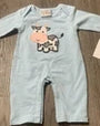A light blue long-sleeve baby romper with a cow graphic on the chest, shown flat on a brown wood floor next to a paper tag.