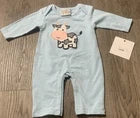 A light blue long-sleeve baby romper with a cow graphic on the chest, shown flat on a brown wood floor next to a paper tag.