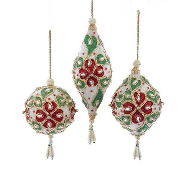 Three Christmas tree ornaments hanging against a white background: one teardrop-shaped ornament in the center and two round ornaments on either side. The ornaments are cream-colored with red and green floral patterns and small bead accents.