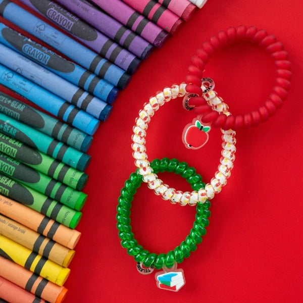 Three beaded stretch bracelets in red, green, and white are arranged on a bright red background next to a row of colorful crayons. Each bracelet has a small holiday-themed charm, including a red apple with a green leaf and a green leaf with a red maple leaf.