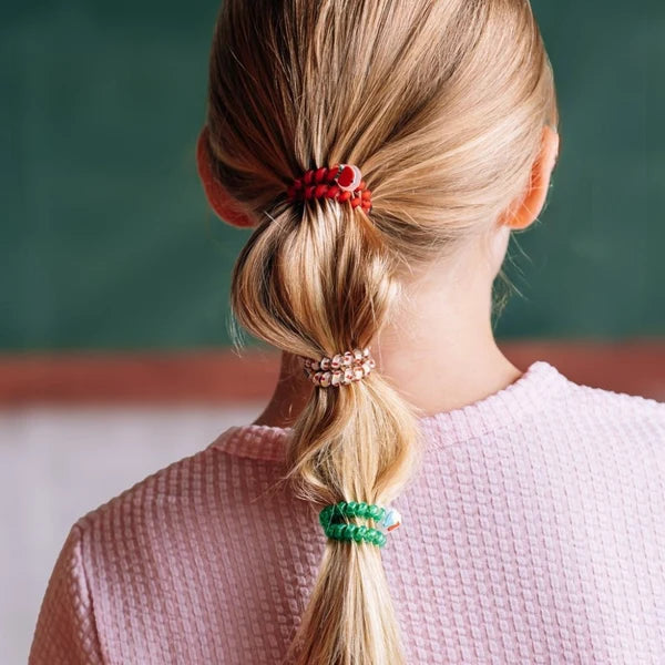 A person shown from the back wearing three colorful hair ties in red, clear rhinestone, and green, with blonde hair styled in a low ponytail. The person is wearing a pink textured sweater against a dark green chalkboard background.