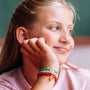 A close-up of a young girl with blonde hair resting her chin on her hand, wearing a set of colorful braided bracelets on her wrist. The bracelets are red and green with small clear charm beads.
