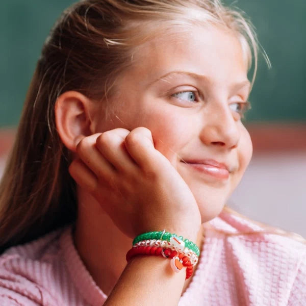 A close-up of a young girl with blonde hair resting her chin on her hand, wearing a set of colorful braided bracelets on her wrist. The bracelets are red and green with small clear charm beads.