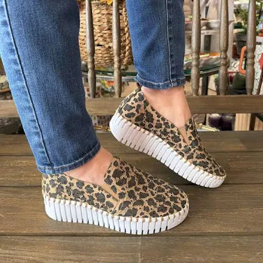 Leopard print slip-on shoes with white soles worn by a person on a wooden floor.