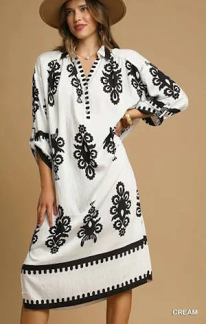 A woman wearing a white long-sleeve tunic dress with black floral embroidery and a black scalloped hem, styled with a brown wide-brim hat against a solid beige background.