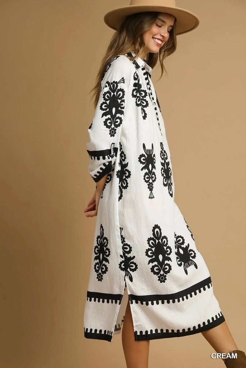 A woman wearing a cream-colored long-sleeve midi dress with black floral embroidery and a black geometric border, paired with a wide-brimmed tan hat, set against a solid beige background.