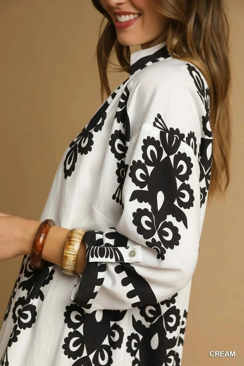 A woman wearing a white long-sleeve tunic-style top with bold black floral and scroll patterns, shown in a close-up side view against a warm beige background.