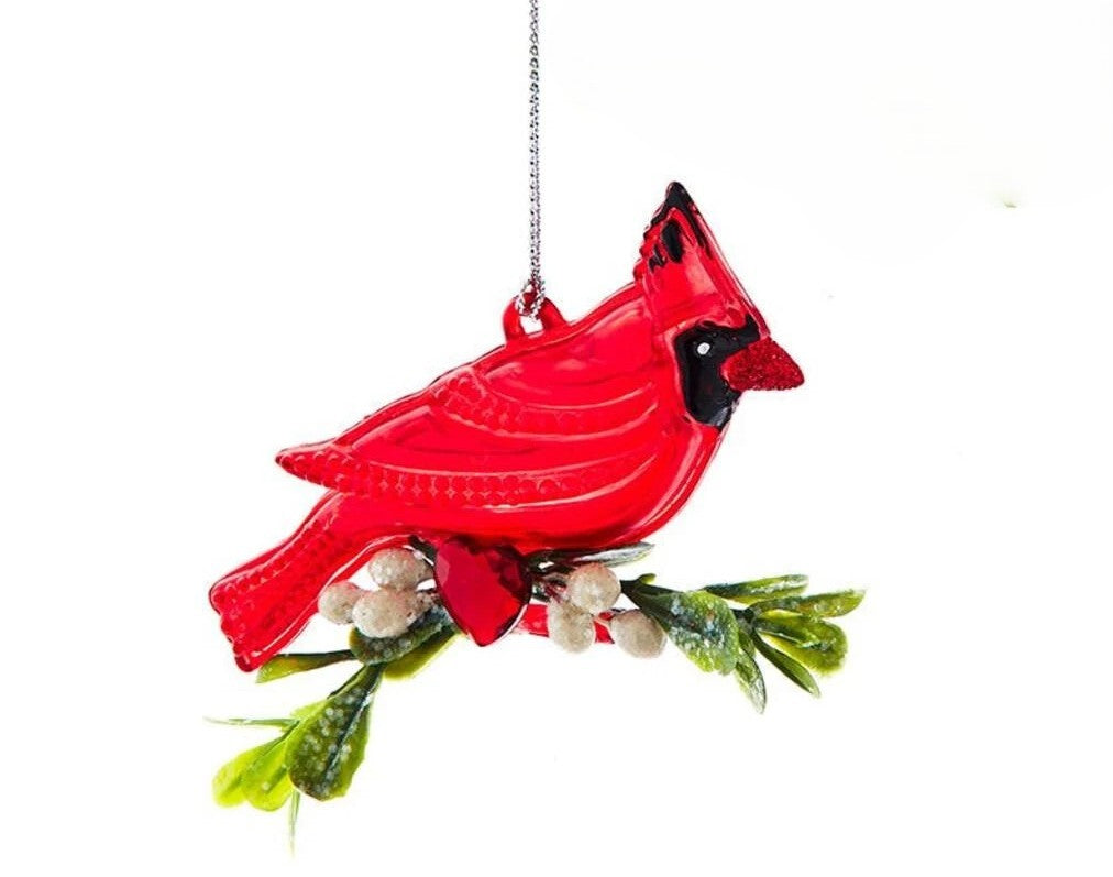 A red cardinal bird hanging ornament perched on a green branch with small white berries, suspended by a silver chain against a plain white background.