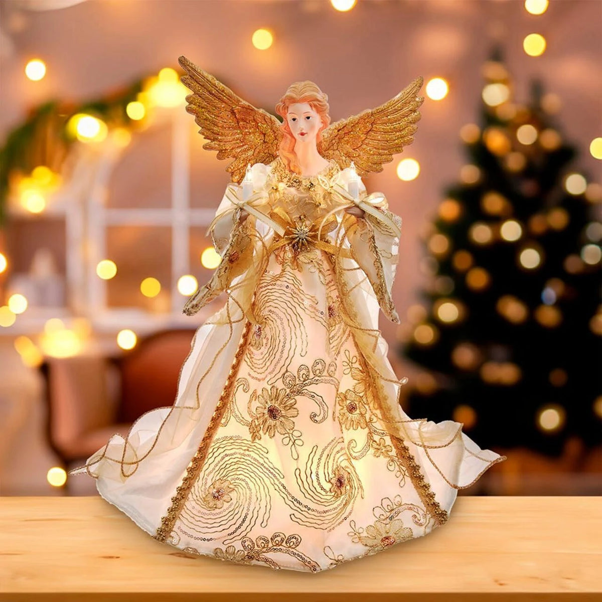 An angel Christmas tree ornament with gold wings and a white gown decorated with gold swirls and floral patterns, displayed on a wooden surface with a blurred Christmas tree and warm lights in the background.