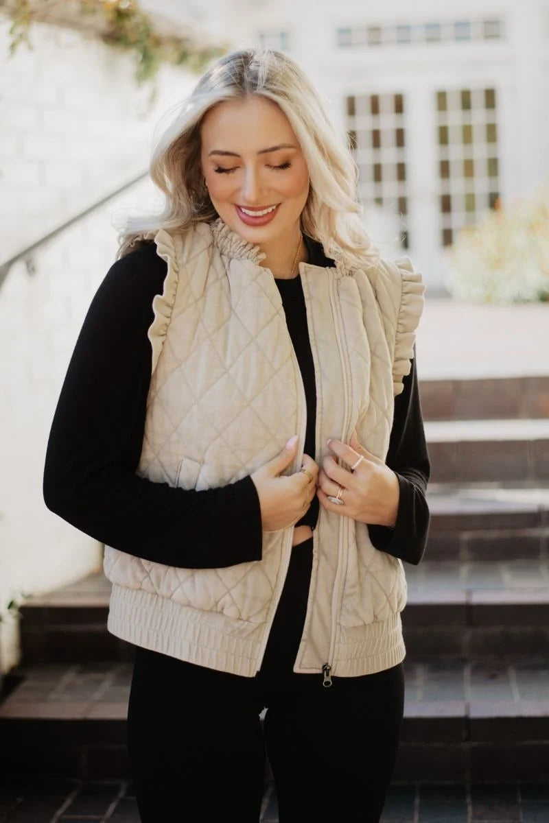 A woman wearing a cream-colored quilted zip-up vest with ruffle trim over a black long-sleeve top, standing outdoors on stone steps.