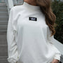 White turtleneck sweater with Lulu Mac logo worn by a person outdoors.
