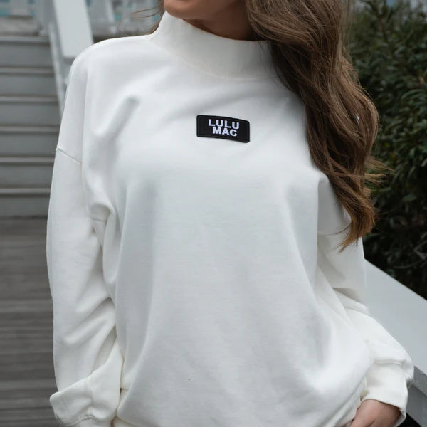 White turtleneck sweater with Lulu Mac logo worn by a person outdoors.