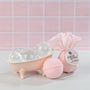 Pink bath bomb with packaging and a small pink bathtub on a light pink tiled background