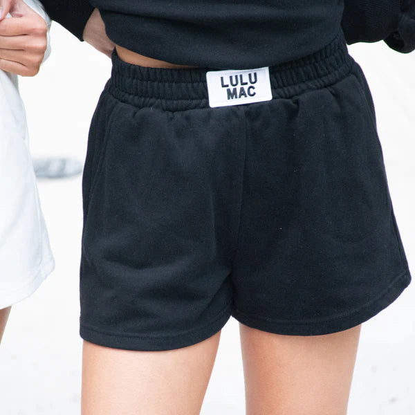 Black shorts with 'LULU MAC' label worn by a person on a white background